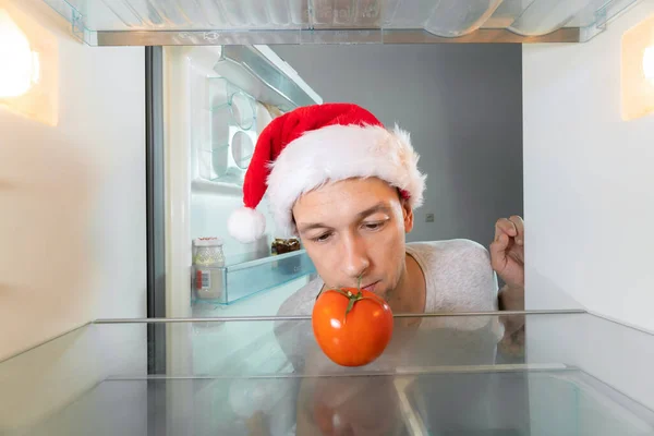 Man in a Santa Claus hat looks suspiciously at a tomato in an empty refrigerator. Concept of an empty refrigerator, delivery service, hunger. Camera inside the refrigerator
