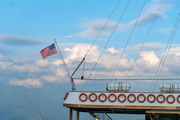 American flag on the mast of the ship against the background of mountains with clouds on a sunny day
