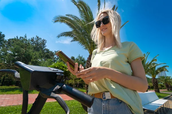 Young happy blonde woman in sunglasses, a light green T-shirt, jeans scans the QR code with a mobile phone to activate the scooter on a sunny day against the background of palm trees