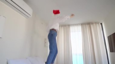 Noel Baba şapkalı güzel beyaz kadın müzik eşliğinde dans ediyor, yatağa atlıyor, Noel tatilinin tadını çıkarıyor otel odasında ya da dairesinde yalnız başına. mutluluk ve neşe.