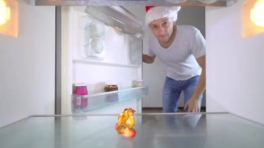 young man in a Santa Claus hat opens refrigerator after party and looks inside, there is an apple core on shelf and an empty refrigerator without food. Hungry time and crisis. dismissal from work.