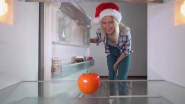 woman in Santa Claus hat opens refrigerator for Christmas, looks inside disappointed, an empty refrigerator. She takes out her smartphone and orders groceries in app. Online order for food delivery.