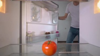 young man in Santa Claus hat opens refrigerator after the party and looks inside, there is a tomato on the shelf and an empty refrigerator without food. Hungry time and crisis. dismissal from work.