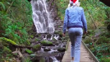 The camera follows a young blonde in a Santa Claus hat through a curved tree, against the background of the Mirveti Waterfall in Georgia. A female traveler walks on Christmas Day. Christmas nature .