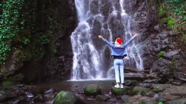 A young blonde in a Santa Claus hat stands on the Mirveti Waterfall in Georgia, arms outstretched. A female traveler celebrates a walk on Christmas Day. Christmas nature. Christmas Eve