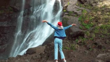 A girl in a Santa Claus hat enjoys nature and dances against the backdrop of a waterfall. A blonde in a denim suit performs a hip dance against the background of the Mahuntseti waterfall Georgia.