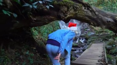 The camera follows a young blonde in a Santa Claus hat through a curved tree, against the background of the Mirveti Waterfall in Georgia. A female traveler walks on Christmas Day. Christmas nature .