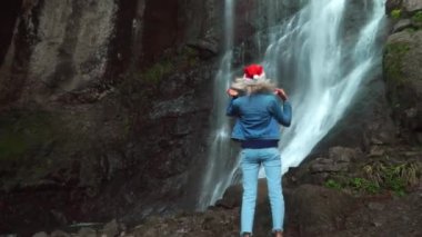 young blonde in Santa Claus hat stands at Makhuntseti waterfall in Georgia with her hair straightened. traveler celebrates walk on Christmas Day. She looks and enjoys nature around fixing her hair.