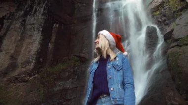 A young blonde in a Santa Claus hat stands at Makhuntseti Waterfall in Georgia. female traveler celebrates a walk on Christmas Day. Christmas nature. Christmas Eve. She looks and enjoys nature around.