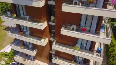Close-up from drone offers view of modern multi-storey building with many windows and balconies. Copter flies around hotel in close-up. flowers and tables with chairs on terrace. Tourist season