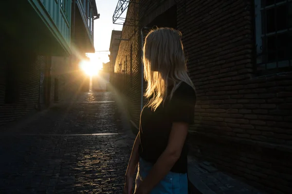 Blonde young woman in a black T-shirt and shorts, standing on a cobblestone street of the old town, turned over her shoulder and looks at the departing sun. Street photography