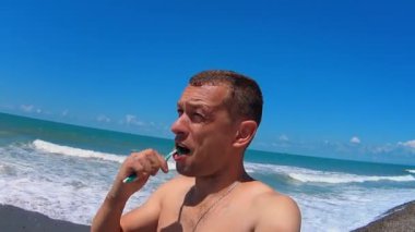 Young man brushes his teeth while standing outside against backdrop of sea. Oral care in extreme conditions. Picnic or camping in nature. Self-care. modern tourism.