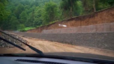 Car is moving along road washed out by rains, camera shoots inside from cabin. Mountain road of clay in rain. Impassable places. brushes clean window. Impassability.