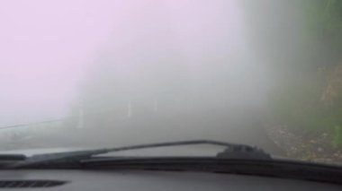 First-person view of driver of car driving down road with thick fog or smog. View from inside car, poor visibility. Countryside, road through forest and mountains.