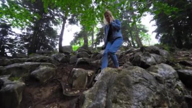 Female tourist walks through wild landscaped forest with trees and moss on rocks. Atmosphere of eerie or fairy-tale forest with large stones and mountains. Fog or smog enveloped everything,