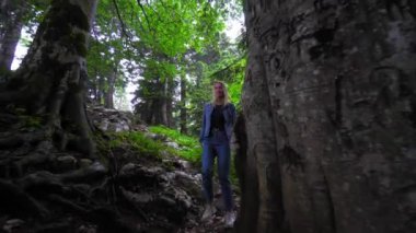 Female tourist walks through wild landscaped forest with trees and moss on rocks. Atmosphere of eerie or fairy-tale forest with large stones and mountains. Fog or smog enveloped everything,