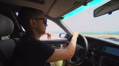 Young Caucasian man drives car on sunny day. He wore black sunglasses on way and stuck his hand out window to catch currents of air and wind. Trip or travel by car.