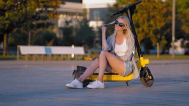 Beautiful young woman in sunglasses sits on her electric scooter in city park and touches her hair while enjoying warm sunny day. Spends weekends. Happy modern people.