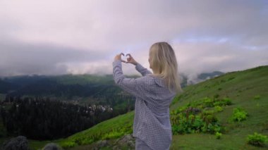 Young girl shows her heart with her fingers against background of clouds in high-altitude village in Georgian district of Bakhmaro. happy, beautiful view of clouds. love for mountains and nature.