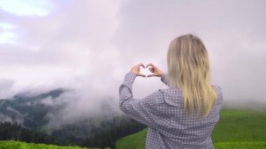 Young girl shows her heart with her fingers against background of clouds in high-altitude village in Georgian district of Bakhmaro. happy, beautiful view of clouds. love for mountains and nature.