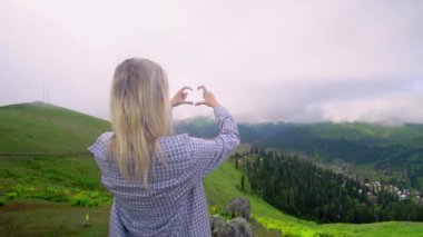 Young girl shows her heart with her fingers against background of clouds in high-altitude village in Georgian district of Bakhmaro. happy, beautiful view of clouds. love for mountains and nature.