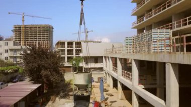 Industrial crane, working and lifting cargo in form of container with concrete. Drone monitors load and rises up. Aerial view. Against background houses under construction and city. Developer