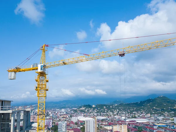 Close-up view from a drone of a yellow construction crane against the background of the city and mountains on a sunny day. Concept of construction, industry, business