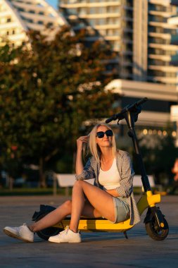 Smiling beautiful young slender blonde woman in denim shorts, T-shirt, shirt, sneakers, sunglasses is sitting on a yellow scooter and enjoying life in a city park at sunset. Vertical photo