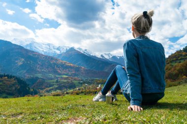 Close-up of a young woman with a cup of coffee sitting on the grass and admiring the beautiful,incredible view of the mountains on a sunny autumn day.Concept of travel,outdoor.