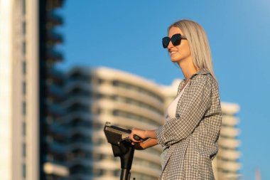 Portrait of a young smiling beautiful blonde woman in a shirt, sunglasses with an electric scooter in her hands on a background of blue sky, buildings on a sunny day. Copy space