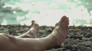 Close-up of women's feet lying on pebble beach against backdrop of sea and waves. Glare of sun is reflected on waves, woman rests and sunbathes. Kind of relaxation and enjoyment in sunbath.