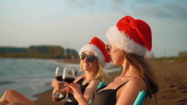 穿着比基尼的快乐女孩们坐在沙滩上庆祝圣诞节的椅子上 戴着桑塔的红帽子喝酒 年轻的微笑的女人 圣诞海滩派对的理念 击中眼镜 新的一年 — 图库视频影像