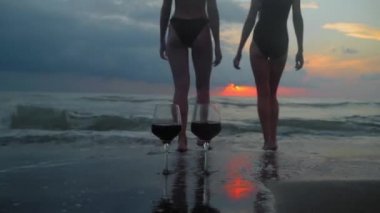 Romantic evening with stunning sunset, two women entering scream at sunset, against backdrop of two glasses of red wine and sea waves. Beautiful landscape on vacation or resort. Pair.
