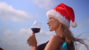 Kış tatili kavramı. Havuzda, siyah mayo ve Noel Baba şapkasıyla mavi gökyüzüne yaslanmış, camdan kırmızı şarap içen güzel bir kız. Okyanusta kış tatili