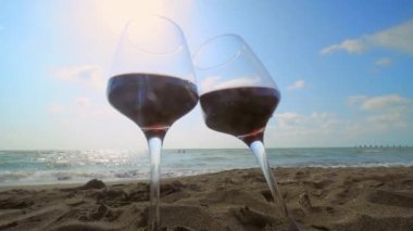 Romantic evening with stunning sunset, two glasses of red wine against backdrop of sea waves. Waves hit glass and sun illuminates wine in glasses. Beautiful landscape on vacation or resort. Pair.