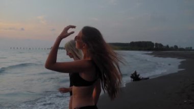 Two young beautiful women in black bikini walking along beach, along sea or ocean. stroll against background of waves and blue sky on horizon. Rest of two friends in sexy clothes. vacation on warm day