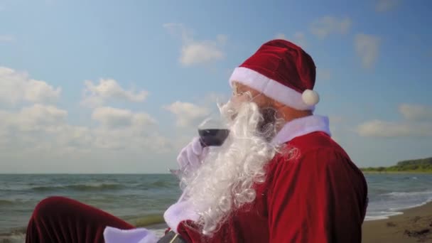衣装を着たサンタクロースは 海や海の近くのビーチでガラスから赤ワインを飲みます 暑い気候の異国の国ではクリスマスを祝う 新年の前に休息と楽しさ リラクゼーションと幸福 — ストック動画