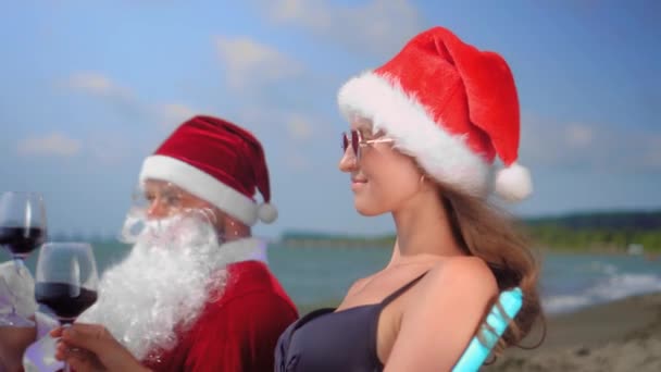 サンタクロースはサンタの帽子のセクシーな女性とのチェイスの長さでリラックスしています 晴れた暑い日には海や海ではなくワインを飲みます サングラス クリスマスと大晦日の夜 — ストック動画