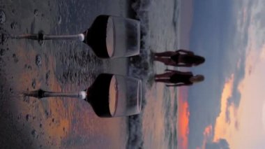 Romantic evening with stunning sunset, two women entering scream at sunset, against backdrop of two glasses of red wine and sea waves. Beautiful landscape on vacation or resort. Pair.