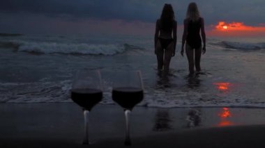 Romantic evening with stunning sunset, two women entering scream at sunset, against backdrop of two glasses of red wine and sea waves. Beautiful landscape on vacation or resort. Pair.