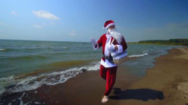 Takım elbiseli Noel Baba deniz kıyısında omzunda torbayla yürüyor. Sahilde, sıcak havada kumlu ve egzotik iklimli bir adada egzotik bir Noel karşılaşması. Dalgalar, deniz. Tatilde dinlenme zamanı..