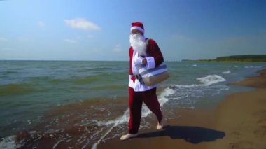 Takım elbiseli Noel Baba deniz kıyısında omzunda torbayla yürüyor. Sahilde, sıcak havada kumlu ve egzotik iklimli bir adada egzotik bir Noel karşılaşması. Dalgalar, deniz. Tatilde dinlenme zamanı..