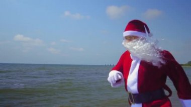 Santa Claus in suit walks and jumps along sea or ocean shore. Exotic Christmas encounter on island on beach with sand in hot weather or exotic climate. Waves, sea. Time to relax on vacation.