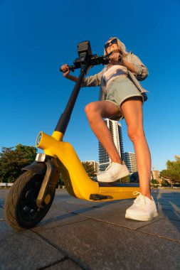 Young slender smiling blonde woman in shorts, shirt, T-shirt, sneakers and sunglasses holds her yellow electric scooter in a city park on a sunny summer evening. Photo from below. Vertical photo