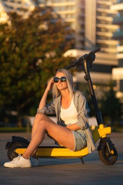 Smiling beautiful young slender blonde woman in denim shorts, T-shirt, shirt, sneakers, sunglasses is sitting on a yellow scooter and enjoying life in a city park at sunset. Vertical photo