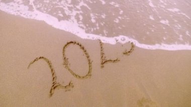 Plajın altın kumu üzerinde 2025 rakamları. Yeni yıl arifesi konsepti. Kıyı şeridinde köpüklü dalgalarla boyanmış odalar. Su dalgası yazıtları temizler. Noel ve Yeni Yıl kavramı.