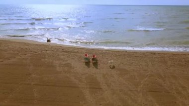 Noel Sahil Tatili Noel Baba Şapkalı Gezgin Kadınlar ve Bikini Tropikal Plajda Kış Tatilinin tadını çıkarın. Kızlar deniz kenarındaki sandalyelere oturur ve şarap kadehlerinin keyfini çıkarırlar. Hava veya İHA saldırısı