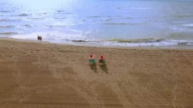İnsansız hava aracıyla çekim yapan, bikinili iki güzel kız Noel Baba şapkalı sandalyelere oturur, şarap içer ve deniz ufkuna ve dalgalara bakarlar. Okyanus kıyısındaki ada. Tropikal ülkede yeni yıl kutlamaları