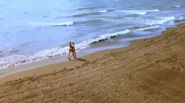 Noel Sahil Tatili Noel Baba Şapkalı ve Bikini 'li Seyahat Kadınları Tropikal plajda kış tatillerinin tadını çıkarın. Kızlar deniz kenarında yürürler ve manzaranın tadını çıkarırlar. Hava veya insansız hava aracı fotoğrafçılığı