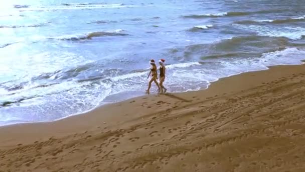 ドローンで撮影すると 2人の美しい女の子がサンタクロースの帽子のビーチを歩き ワインを飲み 海の地平線と波を見ています 熱帯の国での大晦日のお祝い — ストック動画